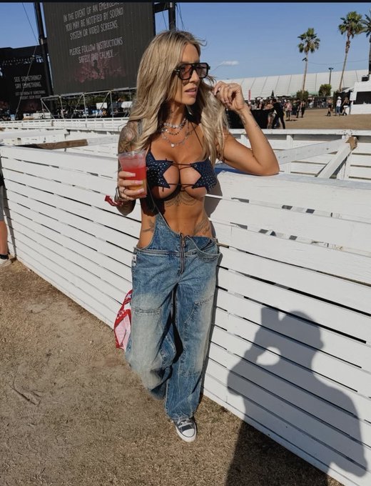 A blonde woman in a polka-dot bikini top, baggy jeans, and sunglasses leans against a white wooden fence holding a pink drink, posing confidently at what appears to be a festival—titled with a tongue-in-cheek caption framing her as the "ugly bitch wife's best friend" to highlight her striking, head-turning festival style.