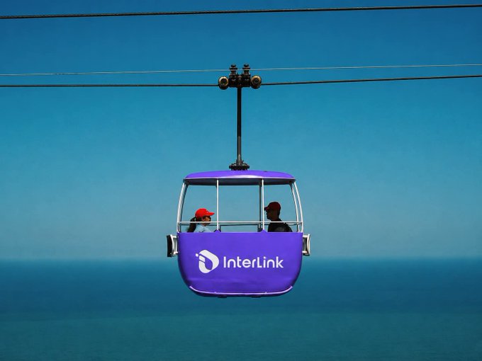 A purple gondola/sky cabin branded with the 'InterLink' logo carrying two passengers across a body of water against a clear blue sky.