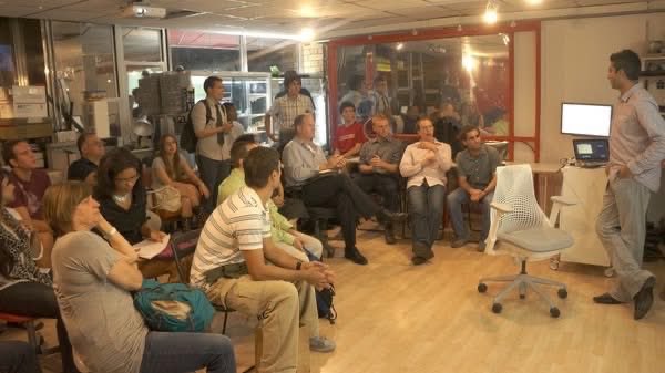 A small group of attendees gathers around a speaker during an intimate Bitcoin meetup in a modest Chinatown venue in 2012, reflecting the grassroots, early-adopter days of cryptocurrency. A monitor displaying what appears to be a Bitcoin-related interface sits at the front, as curious newcomers listen intently about the emerging concept of decentralized money.