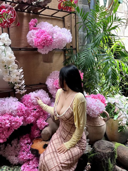 A young woman in a yellow cardigan and pink plaid dress poses among abundant pink and white hydrangea flowers and tropical plants.