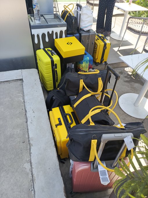 The image shows a collection of luggage, including suitcases and backpacks, stacked up against a wall or ledge outside an establishment. The luggage appears to be packed for travel, with various sizes and colors. There are at least six suitcases visible in the scene, along with two backpacks. A person can also be seen standing nearby, possibly waiting for their turn to check into the hotel or simply observing the luggage arrangement.