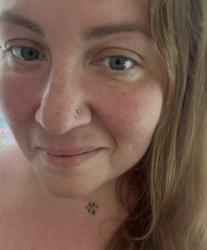 A close-up selfie of a young woman with blue eyes, a nose piercing, and a small floral tattoo on her upper chest.