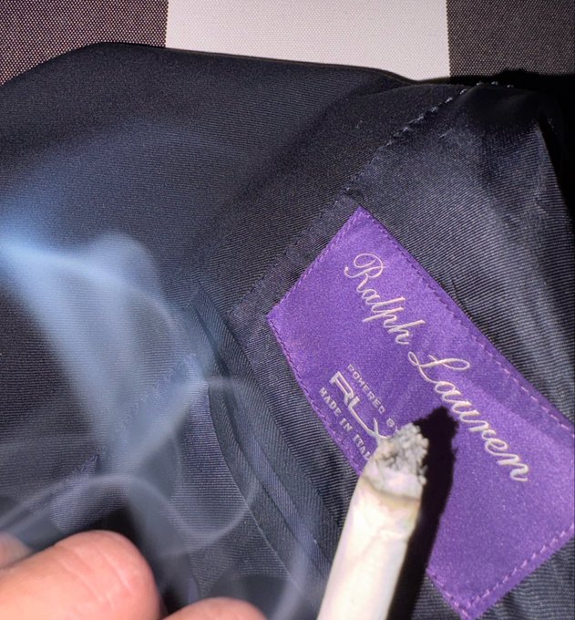 The image shows a person holding a lit cigarette, with smoke coming out of it. They are also smoking a cigarette while looking at an item that has the word "Ralph Lauren" written on it. The background appears to be a black and white checkered patterned fabric or a similar design.