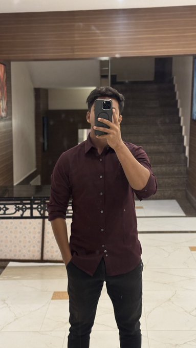 A man takes a mirror selfie in what appears to be a building lobby with a staircase in the background, wearing a maroon button-up shirt and black pants while holding his phone.