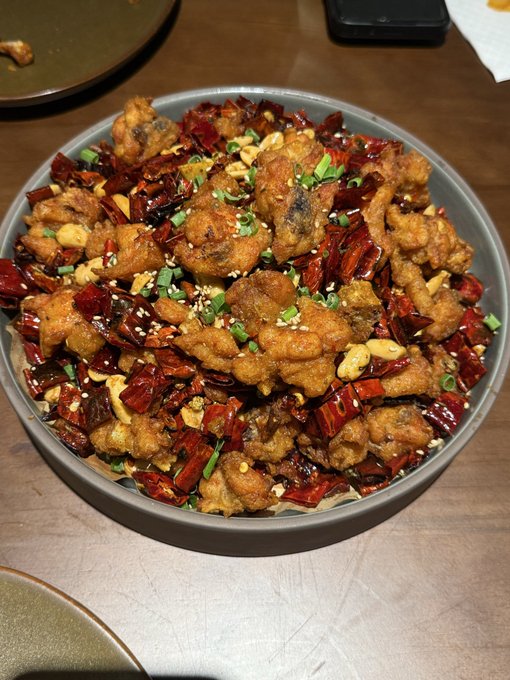 A large gray bowl filled with a Sichuan-style dish of fried chicken pieces mixed with dried red chili peppers, sesame seeds, and chopped green onions, served on a wooden table.