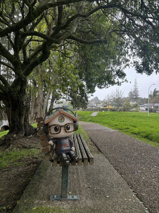 A small figurine with headphones, glasses, and a rainbow badge sits on a wooden bench under a large tree in a park, representing the @PonsGuyNFT team taking a quiet moment in nature before announcing their new website and special surprises for holders today.