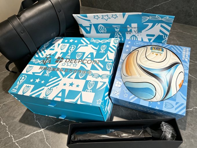 Two DeepCoin-branded boxes (one labeled "DEEPCOIN" in English and Chinese) sit open on a marble surface, revealing a signed soccer ball, a water bottle in bubble wrap, and an envelope with matching blue star-patterned packaging, alongside a black backpack. The image showcases the promotional merchandise from DeepCoin referenced in the post, displaying the quality swag items the Argentine partner sent.