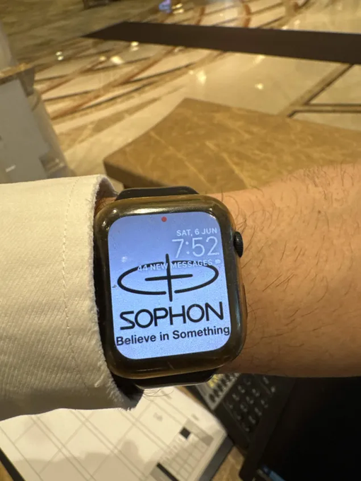 The image shows a person wearing a watch with a screen displaying the word "Sophon" along with some text. The watch has a black band, and the time displayed on the screen reads 7:52 PM. The background of the image is blurred, but there are some chairs visible in the scene.