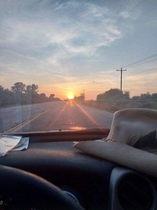 A weathered cowboy hat rests on the dashboard of a truck driving down an open road at sunrise, symbolizing the long journey and dedication behind the speaker's creative process.
