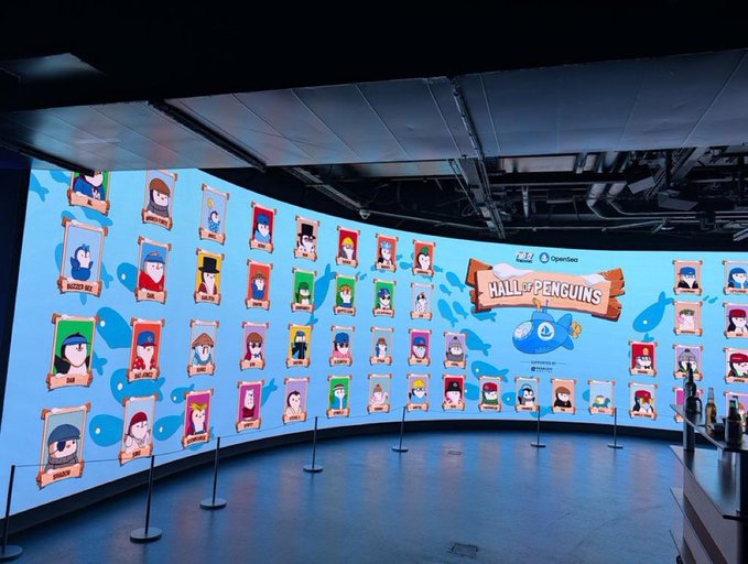 A large curved screen at an event displays 'HALL OF PENGUINS' by Pudgy Penguins and OpenSea, featuring numerous penguin character portraits arranged in a grid.