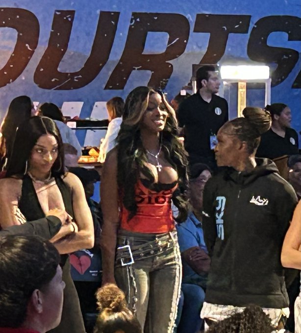 Three women stand courtside at a Chicago Sky event, with the word "COURTS" visible on a blue backdrop behind them. The caption "Timeout in #skytown @chicagosky" aligns with the setting, suggesting they are attending a Chicago Sky WNBA event during a game break, with one woman wearing a red top and cross necklace, another in a black halter outfit, and a third in a black hoodie with stylized lettering.