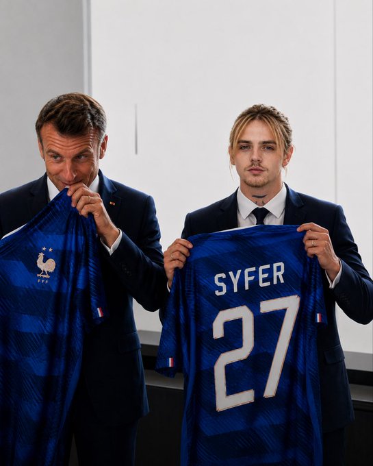 Two men in dark suits hold up a blue French national football team jersey with the name "SYFER" and number "27" printed on the back, standing in front of a white projection screen.