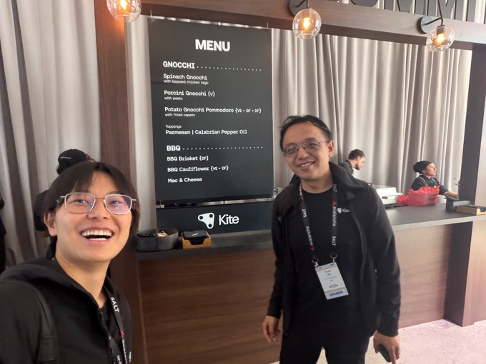 Two smiling attendees pose for a selfie in front of a menu board at a Kite-branded event booth, with the menu listing items like Spinach Gnocchi, Potato Gnocchi Pomodoro, Pastrami Calabrian Pepper, and BBQ options including Brisket and Cauliflower. The scene captures the networking atmosphere referenced in the post about Avalanche Summit and Arc's Mainnet launch in New York, where the team engaged with others at the crypto conference.