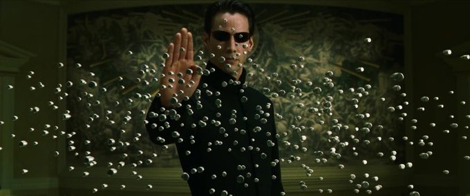 A scene from a film shows a man in a dark suit and sunglasses (Keanu Reeves as Neo) holding up his hand as a swarm of metallic bullet-like objects freezes mid-air in front of him.