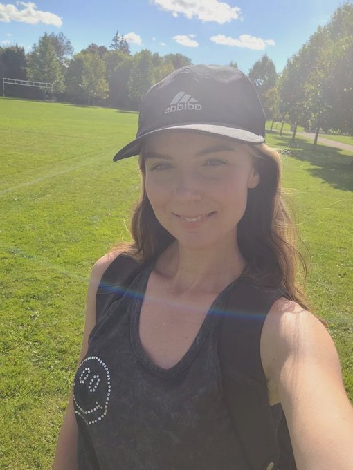 A selfie of a smiling woman wearing a black Adidas baseball cap and a dark grey tank top, standing on a grassy field with trees in the background on a sunny day.