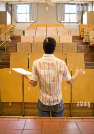 A stock photo showing the back of a person wearing a striped shirt and cap, standing in an empty university lecture hall with wooden desks, holding a notebook as if addressing the class.