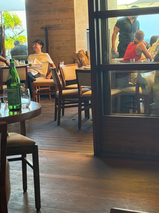 A dimly lit restaurant interior where patrons are seated at wooden tables, with a S.Pellegrino bottle visible in the foreground and staff serving customers through a glass door. The casual dining atmosphere pairs with the "$Kimchi" post, suggesting a relaxed meal at a Korean or Asian-fusion eatery.