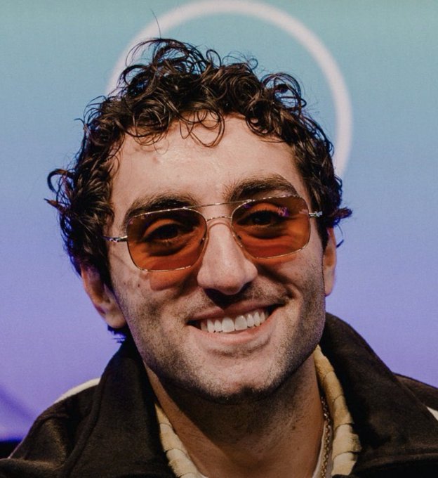A portrait of a smiling man with curly dark hair wearing orange-tinted aviator sunglasses and a brown jacket with a shearling collar, set against a soft blue and purple background with a circular light.