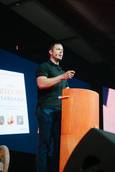 The image features a man standing behind a podium, giving a speech or presentation. The main focus of the image is the speaker, who appears to be wearing a black shirt and blue jeans. There are two chairs visible in the background, one near the left edge of the frame and another closer to the center. A microphone can also be seen on the podium, indicating that this might be an event where he is addressing an audience or delivering a speech. The image does not provide any specific information about the content of the presentation or the post shared in the caption.