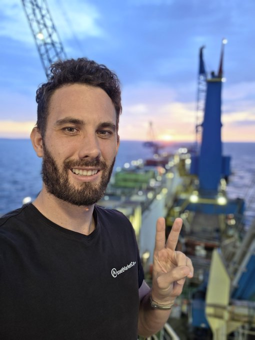 A smiling bearded man wearing a black 'OverthetopCunts' t-shirt takes a selfie making a peace sign, standing on an offshore oil rig or vessel at sunset.