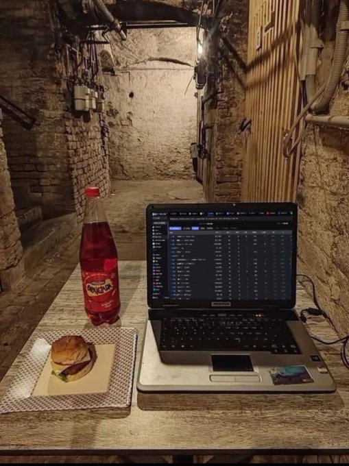 A laptop displaying trading charts sits on a rustic wooden table alongside a bottle of Tuborg soda and a sandwich, set against a dimly lit, stone-walled basement corridor—visually reinforcing the caption's message that fortune is built in humble, unglamorous surroundings.