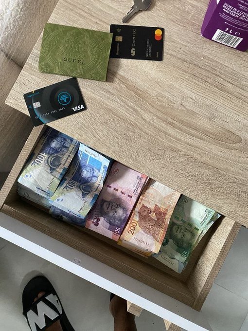 An overhead view shows an open drawer containing stacks of Malaysian Ringgit banknotes in various denominations, along with two credit cards (one Visa, one Mastercard) and a green Gucci card holder on the surface above.