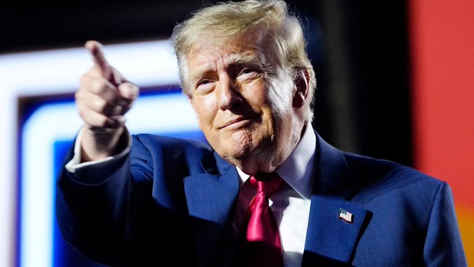 Former President Donald Trump, wearing a navy blue suit with a red tie and American flag pin, points directly at the camera during a rally event. The image accompanies a social media post listing several recent Trump announcements regarding foreign policy, oil prices, legislation, and a proposed $5,000 payment to U.S. citizens, with the post appearing to question the plausibility of these claims.