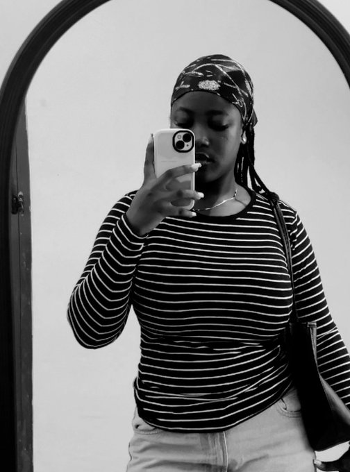 A black and white mirror selfie of a woman wearing a striped long-sleeve shirt and a headwrap, taking a photo with her phone.