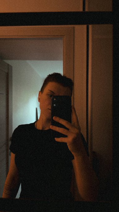 A dimly lit mirror selfie of a person with short hair wearing a black t-shirt, holding a smartphone that partially obscures their face.