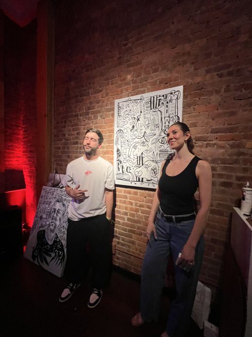 A man in a white t-shirt and a woman in a black tank top stand smiling in front of a brick wall displaying black and white line drawings in an art gallery setting.