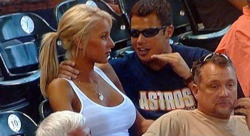 A blonde woman in a white tank top sits next to a man wearing sunglasses and an Astros shirt in stadium seating.