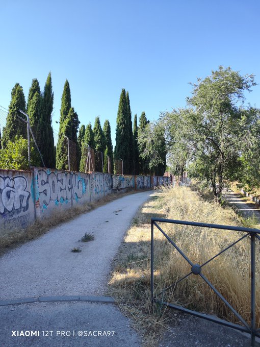 A scenic outdoor photograph of a dirt path bordered by a graffiti-covered wall and tall cypress trees on the left, with dry golden grass and a metal gate on the right under a clear blue sky, watermarked 'XIAOMI 12T PRO | @SACRA97.'