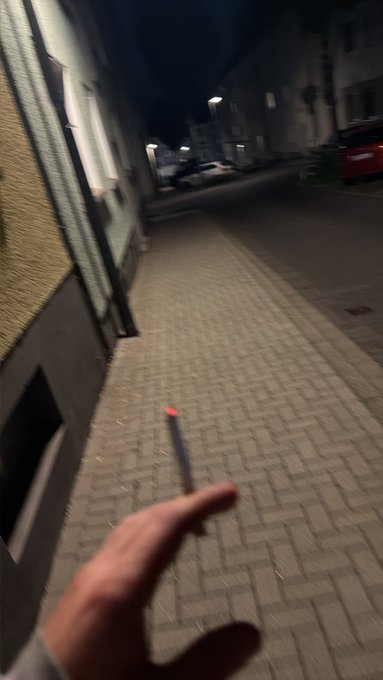 A blurry nighttime scene shows a hand in the foreground near a small red light on a brick-paved street beside a building.