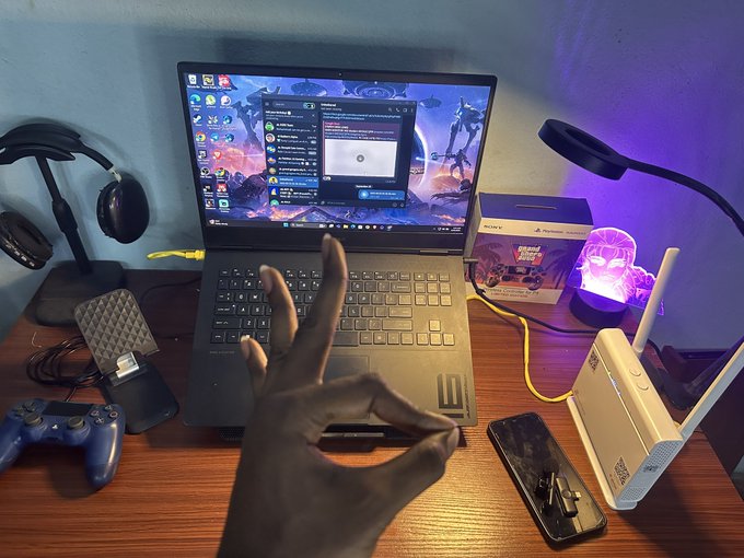 A first-person view of a hand making an OK gesture in front of a laptop on a desk, with a game controller, phone, and router visible in the setup.