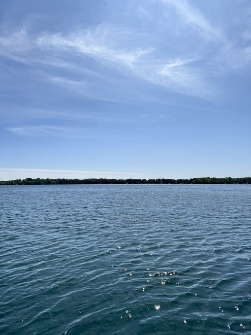 A serene landscape photo shows a calm body of water under a bright blue sky with wispy clouds, with a distant tree-lined shore on the horizon.