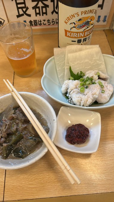 A Japanese meal set on a wooden table featuring a bowl of beef stew, a plate of fish with tofu, a dish of red paste, and a Kirin beer bottle.