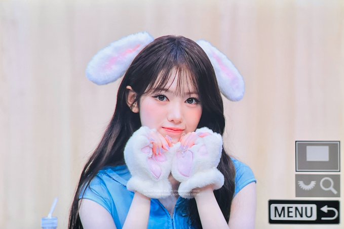 A young woman wearing fluffy white and pink bunny ear headbands and matching paw gloves poses against a soft beige background, with a 'MENU' button visible in the corner.