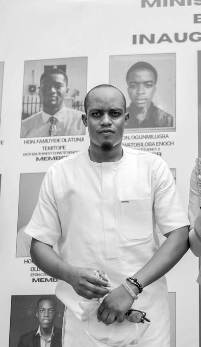 Black and white portrait of a man in white traditional attire standing in front of a backdrop displaying memorial photos and text including 'HON. FAMILYIDE OLATUNJI TEMITOPE', 'ON. OGUNMILUGBA', and 'INAUG'.