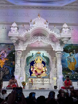 An ornate Hindu temple shrine featuring a statue of Lord Ganesha in white and gold decorative setting with musicians performing in front, while audience members take photos with phones.
