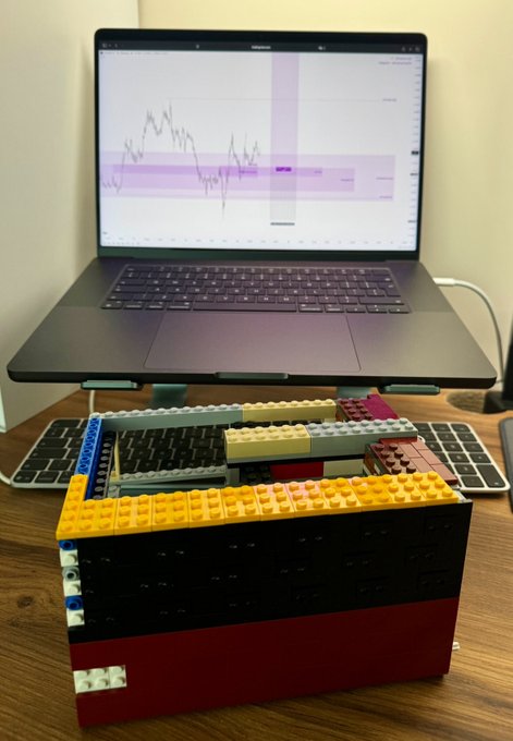 A trader greets the morning ("GM lads!") in front of a laptop displaying a market chart, alongside a LEGO-built rig holding a keyboard elevated above the desk. The setup reflects the casual, meme-friendly tone of the post, which comments on quiet pre-market activity ("PA is so dead") while awaiting the PPI release.