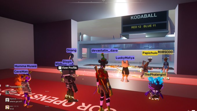 A virtual reality scene with multiple avatars on a red carpet, with the text 'KODABALL RED 12 BLUE 11' displayed on a screen and various usernames floating above the avatars.