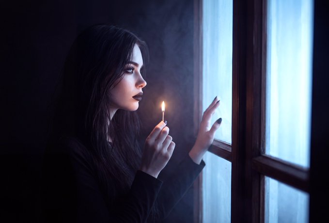 A woman with long dark hair stands by a window in a dark room, lighting a match and illuminating her face.