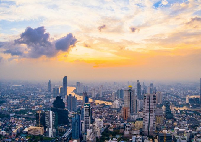 An aerial cityscape at sunset featuring a wide river winding through skyscrapers and dense urban development under a golden-orange cloudy sky.