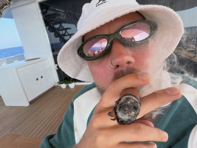 A man with a goatee wearing sunglasses and a white bucket hat while smoking a large cigar on a boat.