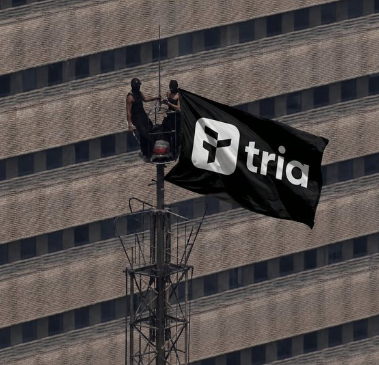 Two people raise a black "tria" flag atop a tower in front of a modern high-rise building, symbolizing the brand's bold presence. The image ties to the post's promotion of the Tria card as a convenient way to spend crypto in everyday transactions.
