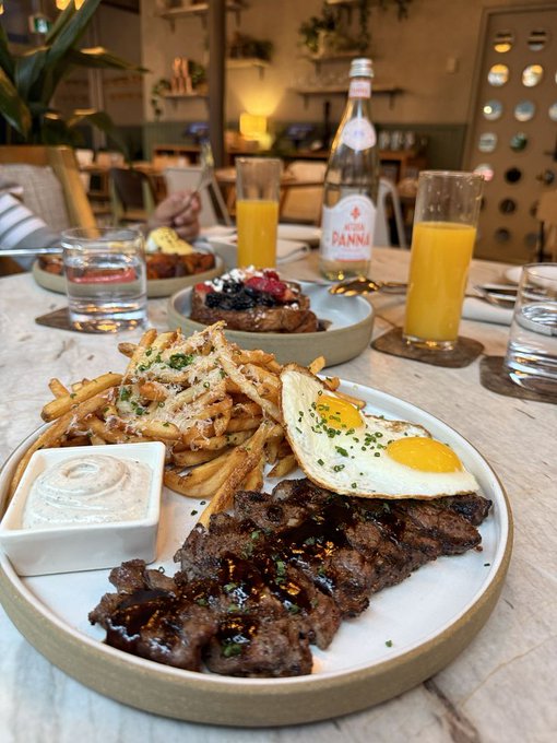 The image shows a dining table set for two people, featuring plates of food such as steak, eggs, french fries, and creme brulee with sauce. There are also glasses of orange juice and water on the table. The scene suggests that this is a casual meal shared between friends or family members at a restaurant.