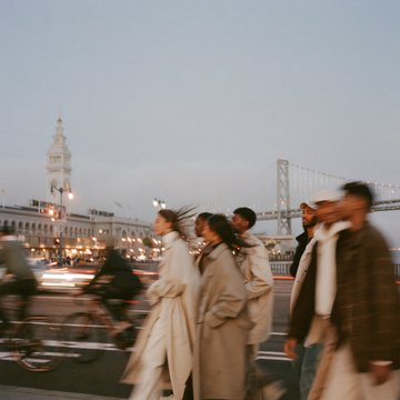 A group of people in fashionable coats walk along the Embarcadero in San Francisco, with the iconic Ferry Building clock tower and Bay Bridge blurred in the background, illustrating the joke about SF needing AI to make the city look cool.