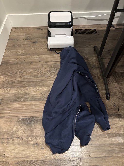 A white and black printer sits on a wood-laminate floor next to a crumpled dark blue jacket or hoodie.