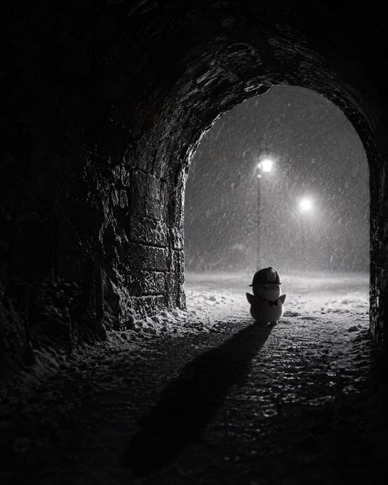 A black and white atmospheric photograph shows a small figure in a hat viewed from behind through a stone archway in snowy conditions.
