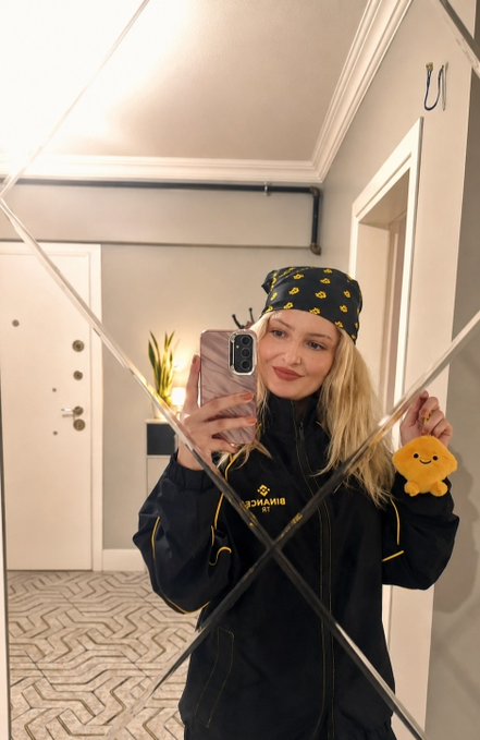 A blonde woman takes a mirror selfie wearing a black jacket with "BINANCE" branding and a matching bandana featuring Binance's yellow logo pattern, while holding a cute yellow plush toy as a gift from BinanceTR. The autumn caption ties in with the cozy setting, showcasing appreciation for the Binance TR promotional gifts.